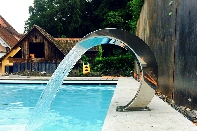 Fontaine cobra en acier inoxydable déversant une lame d'eau dans une piscine, fixée sur une margelle en pierre.