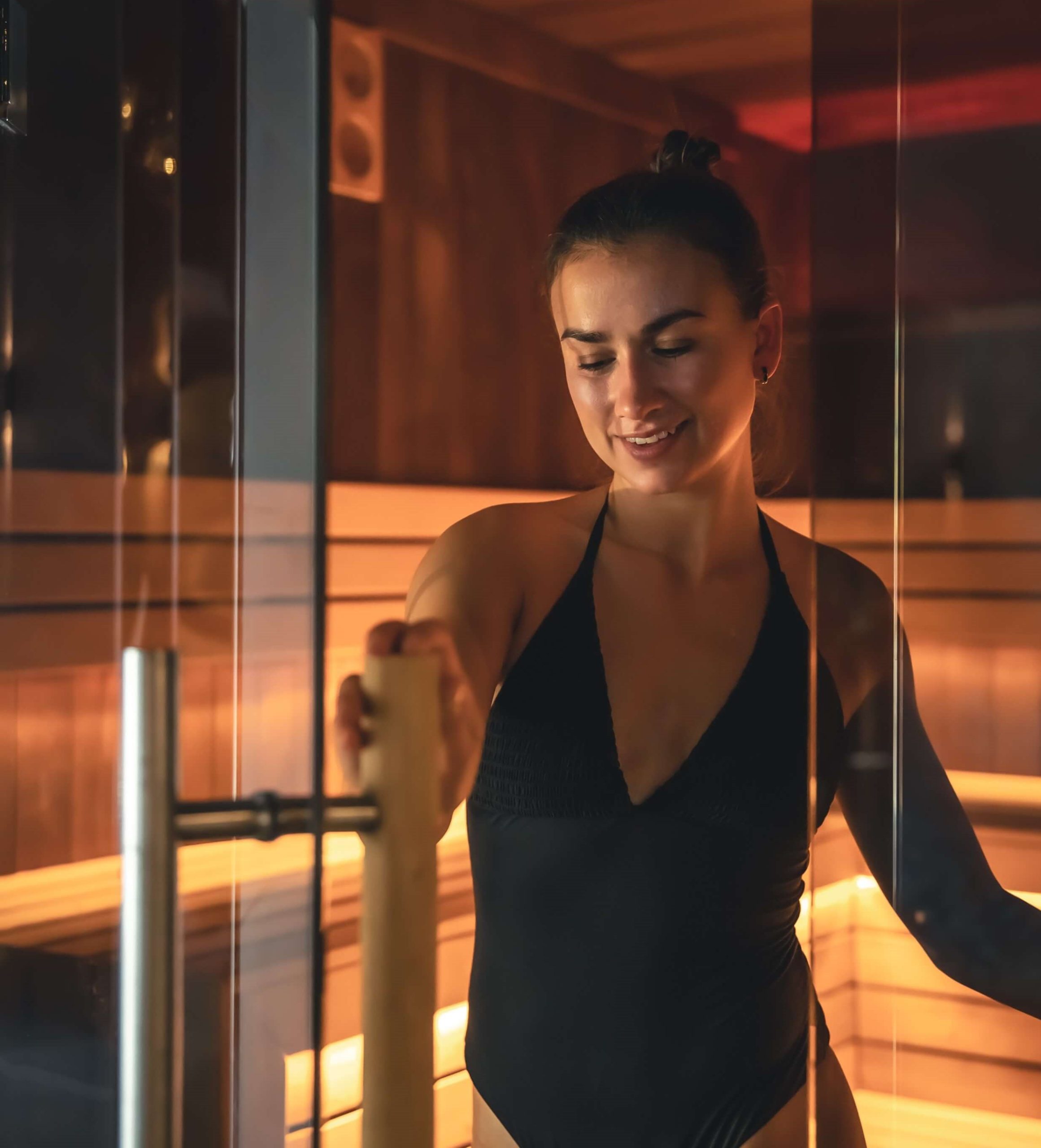 Femme ouvrant la porte d’un sauna contemporain – Pool68 Femme en maillot ouvrant la porte vitrée d’un sauna en bois clair éclairé par des LEDs ambrées.
