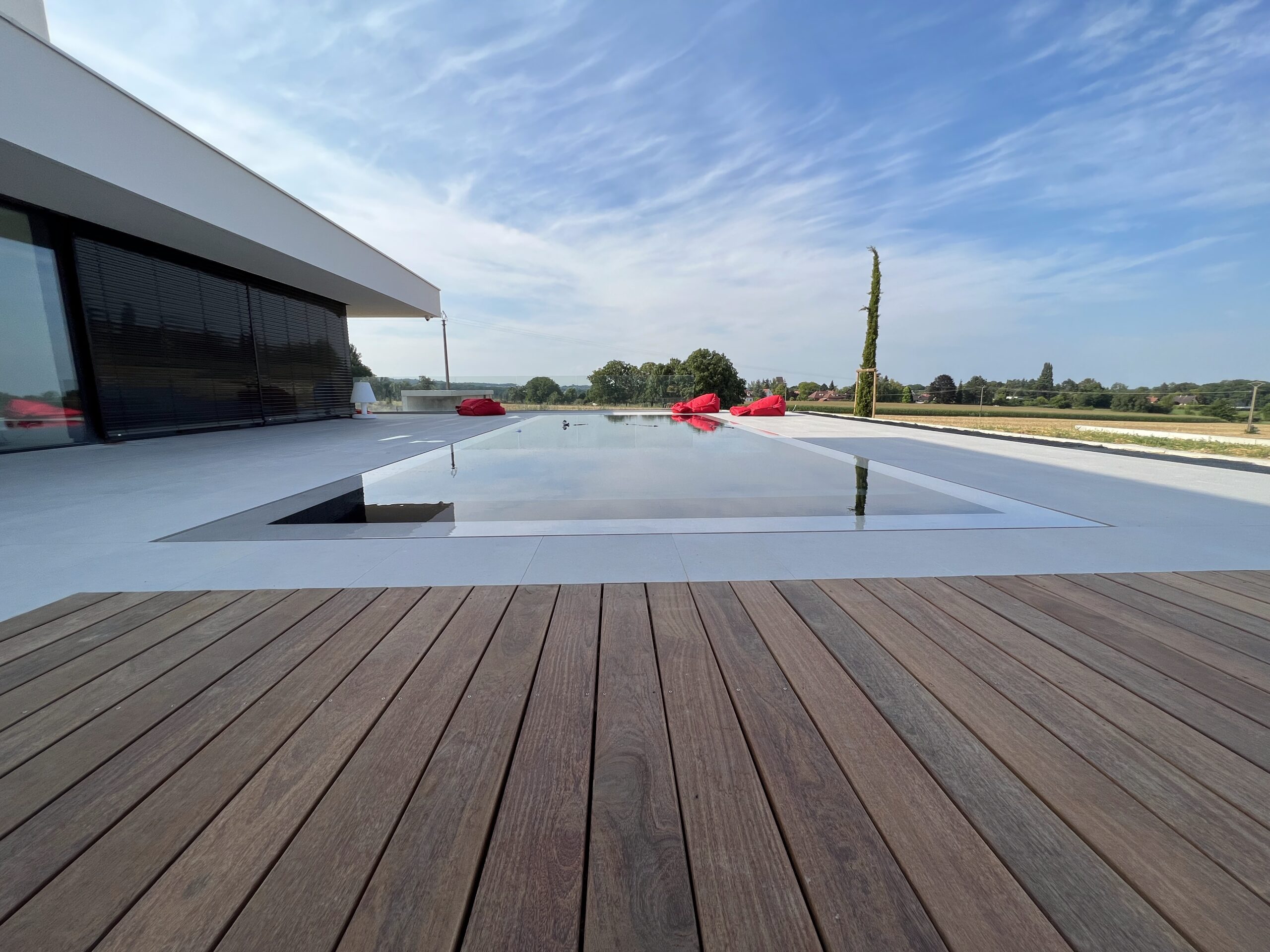 Piscine miroir contemporaine et poufs rouges – Pool68 Longue piscine miroir au liner sombre, eau au ras des dalles grises, terrasse bois, poufs rouges et vue ouverte sur la campagne.