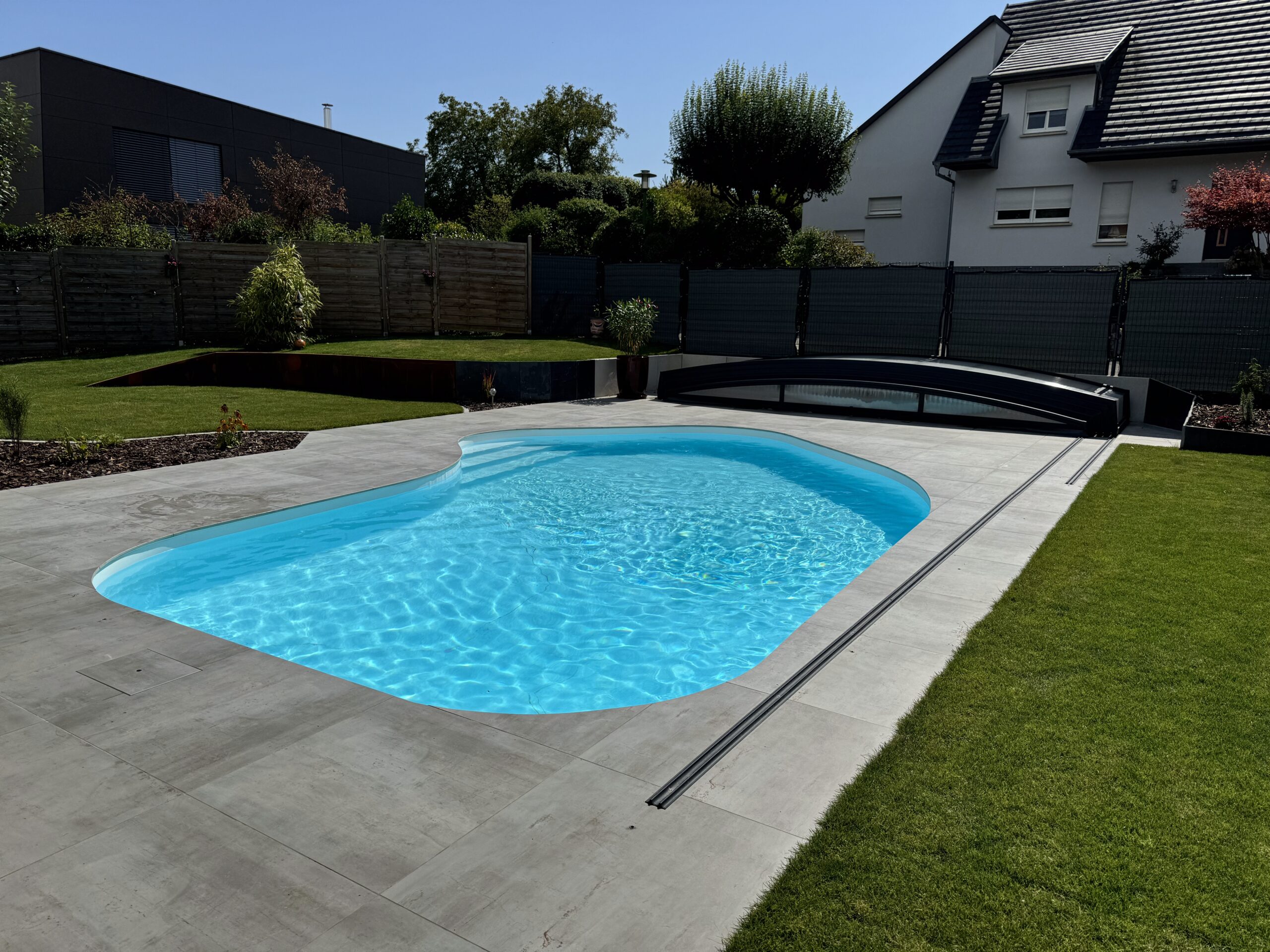 Piscine coque aux formes courbes et abri bas – Pool68 Piscine coque aux formes organiques, eau turquoise, abri télescopique bas anthracite et terrasse en dalles claires.