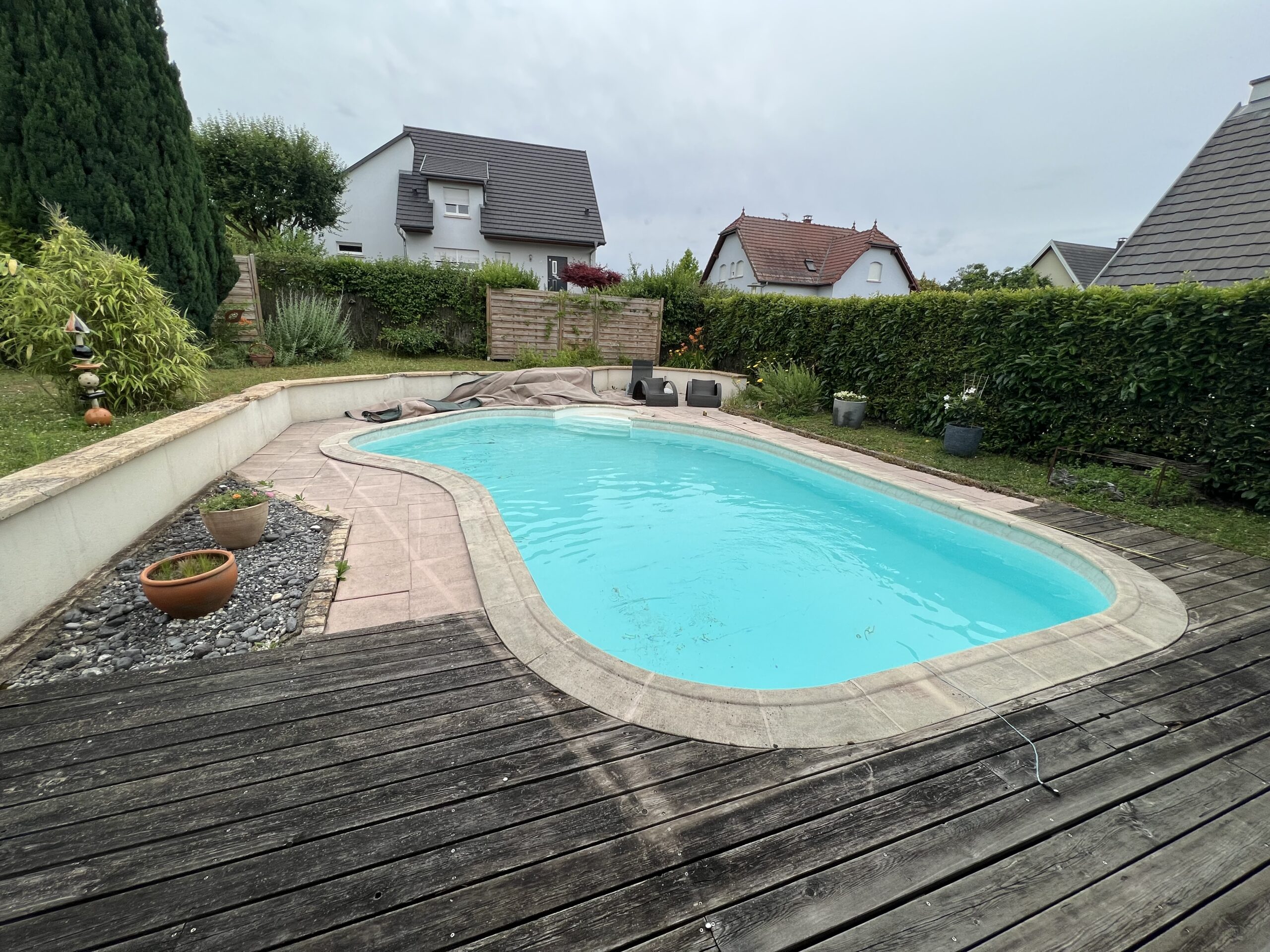 Piscine coque forme haricot et terrasse bois – Pool68 Piscine coque bleu clair en forme de haricot, marches intégrées, terrasse bois, margelles claires et transats dans un jardin.