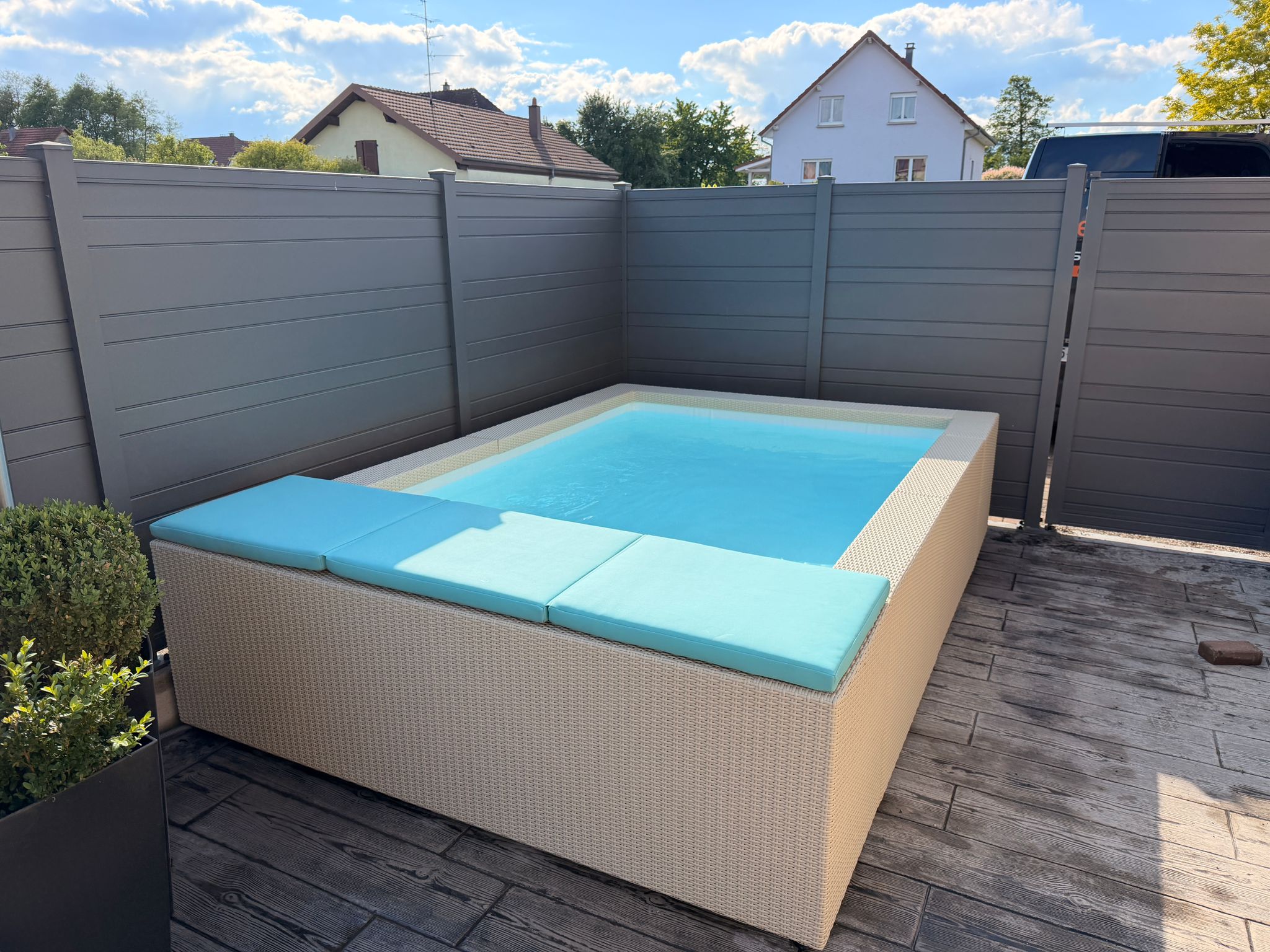Mini-piscine hors-sol habillée en résine tressée – Pool68 Petite piscine hors-sol rectangulaire à habillage résine tressée beige, coussins turquoise, sur terrasse imitation bois.