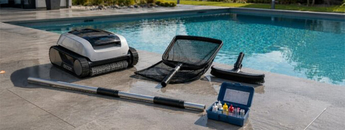 Robot de piscine, épuisette, brosse, perche et mallette d’analyse d’eau au bord du bassin