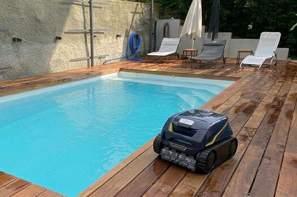 Robot nettoyeur bleu et jaune à chenilles posé sur une terrasse en bois, au bord d'une piscine rectangulaire bleu clair.