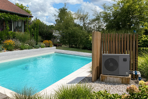 Pompe à chaleur grise installée sur un socle près d'une piscine rectangulaire turquoise, gravier décoratif et jardin.