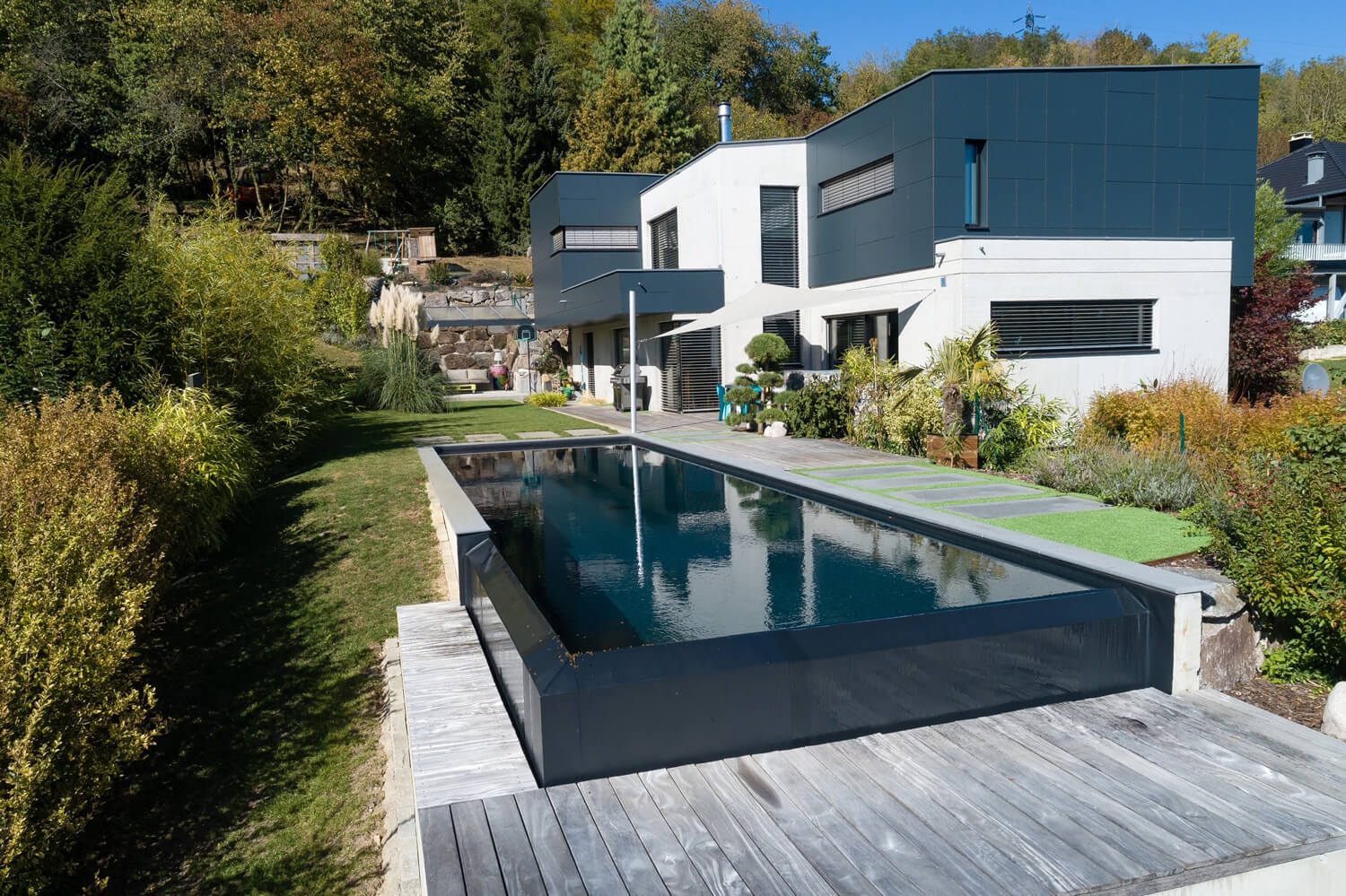 Piscine à débordement et villa contemporaine – Pool68 Piscine miroir à débordement, terrasse bois, pelouse et maison contemporaine au flanc de colline